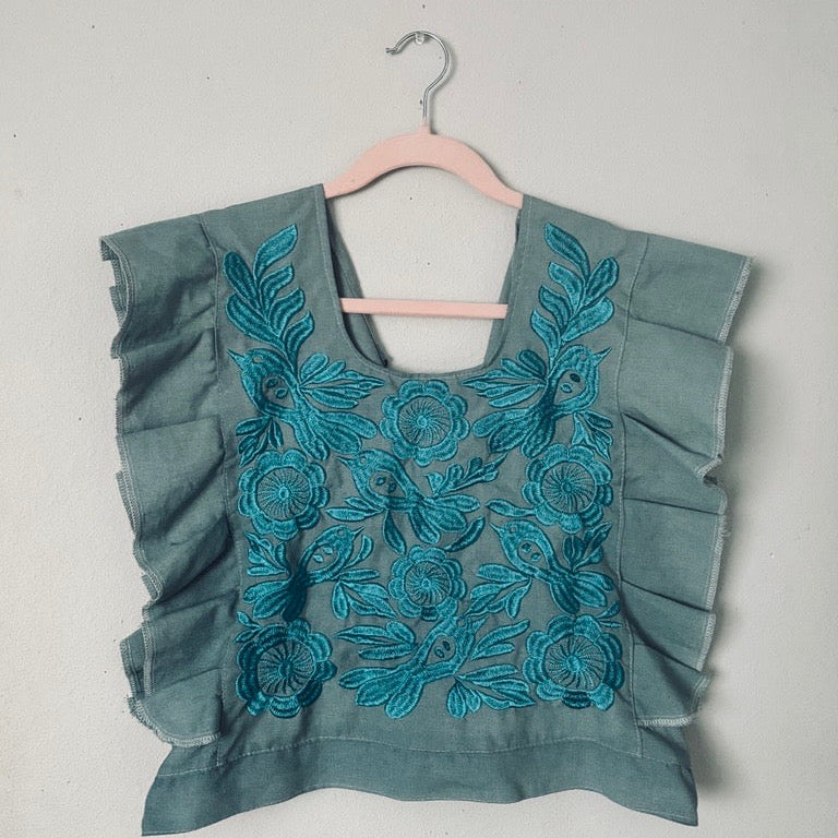 Mexican Teal blouse with floral embroidery on a hanger against a gray background